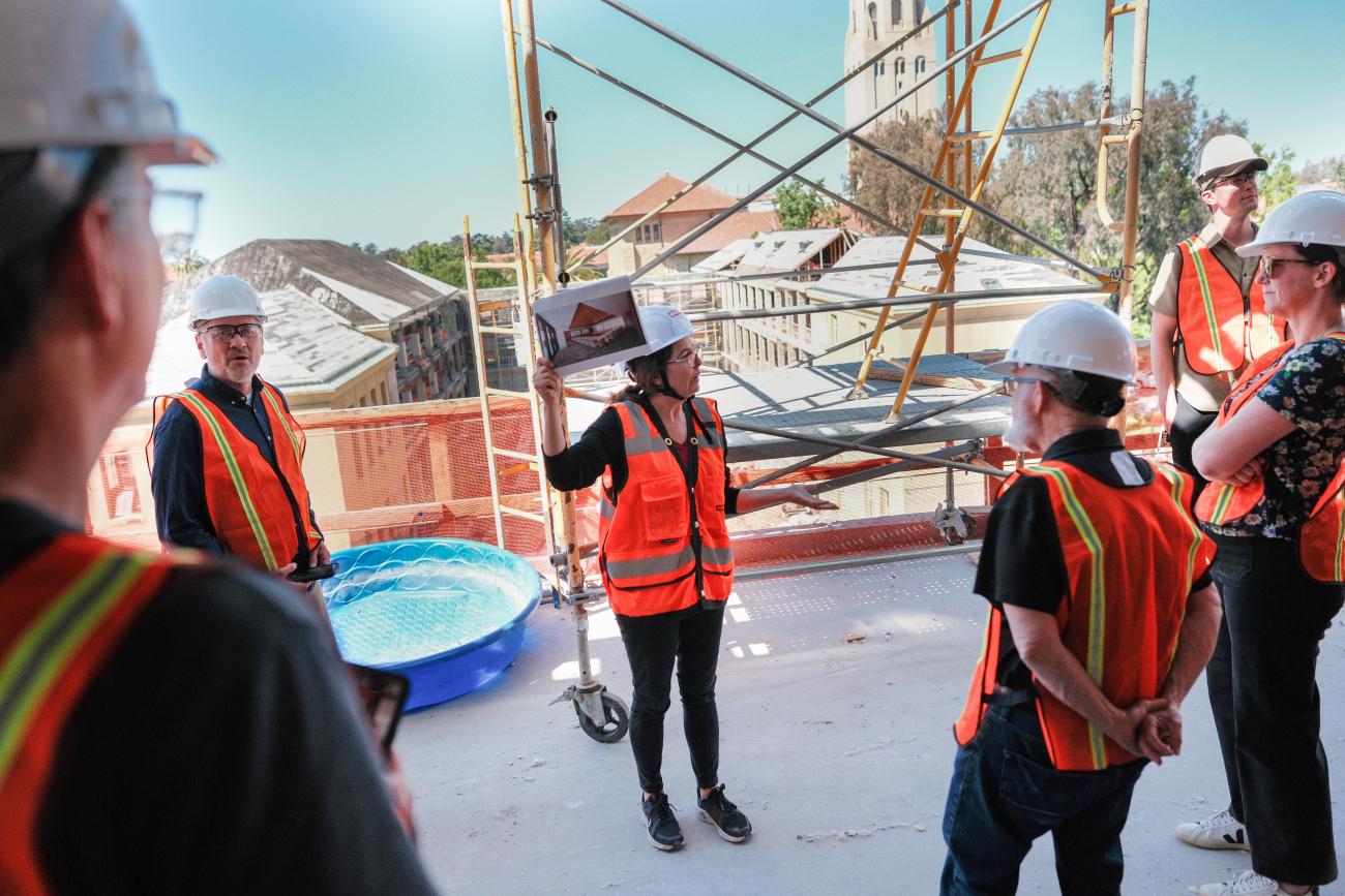 Photo of a group of people, all wearing bright orange construction vests and hard hats, on the top floor of a tall construction site with a view of the old education building and Hoover Tower in the background